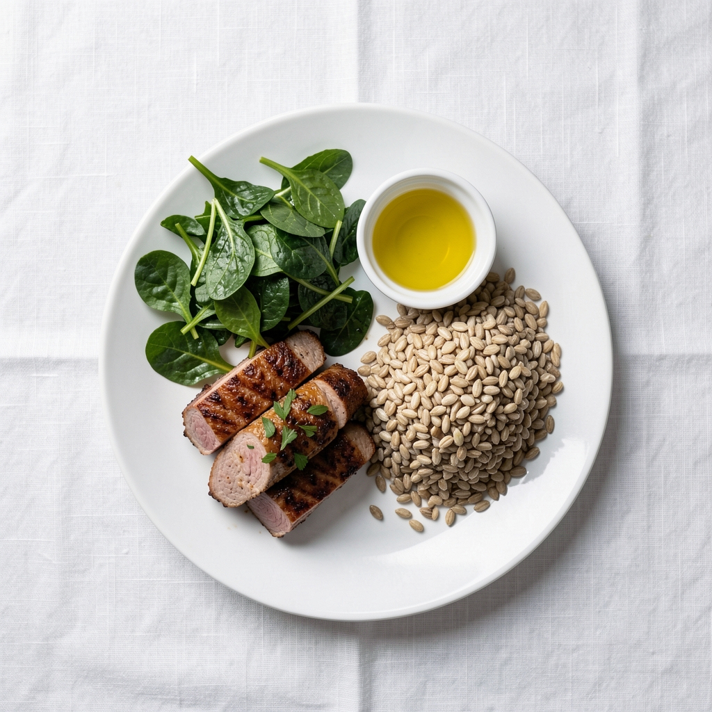 Balanced dinner plate photographed from above on a white linen tablecloth, showing distinct sections of protein, leafy greens, and whole grains with a small ramekin of olive oil
